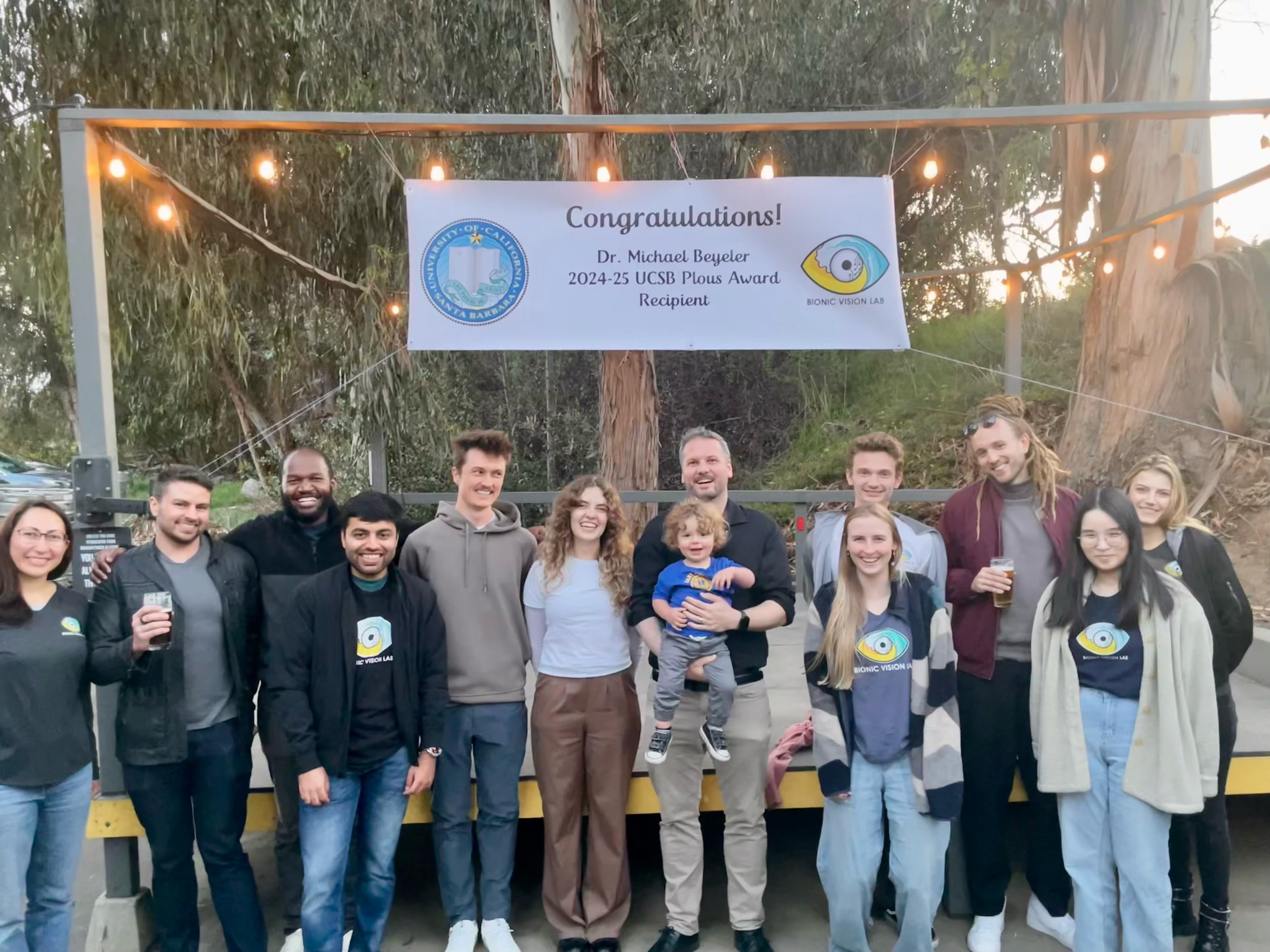 About a dozen lab members, several in Bionic Vision Lab T-shirts, pose under string lights and a banner reading “Congratulations! Dr. Michael Beyeler, 2024-25 UCSB Plous Award Recipient.” From left to right: Roksana Sadeghi, Justin Kasowski, Byron Johnson, Apurv Varshney, Galen Pogoncheff, Lily Turkstra, Prof. Beyeler with Julian, Emily Joyce, Jacob Granley, Marius Schneider, Yuchen Hou, and Hannah Stone.