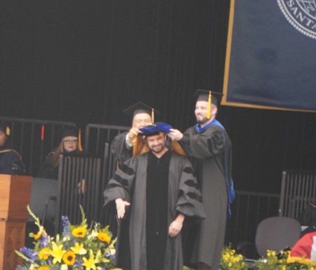 On the commencement stage, Profs. El Abbadi and Beyeler in academic regalia place a doctoral hood over Justin Kasowski’s shoulders, the first graduate from the Bionic Vision Lab (2023).