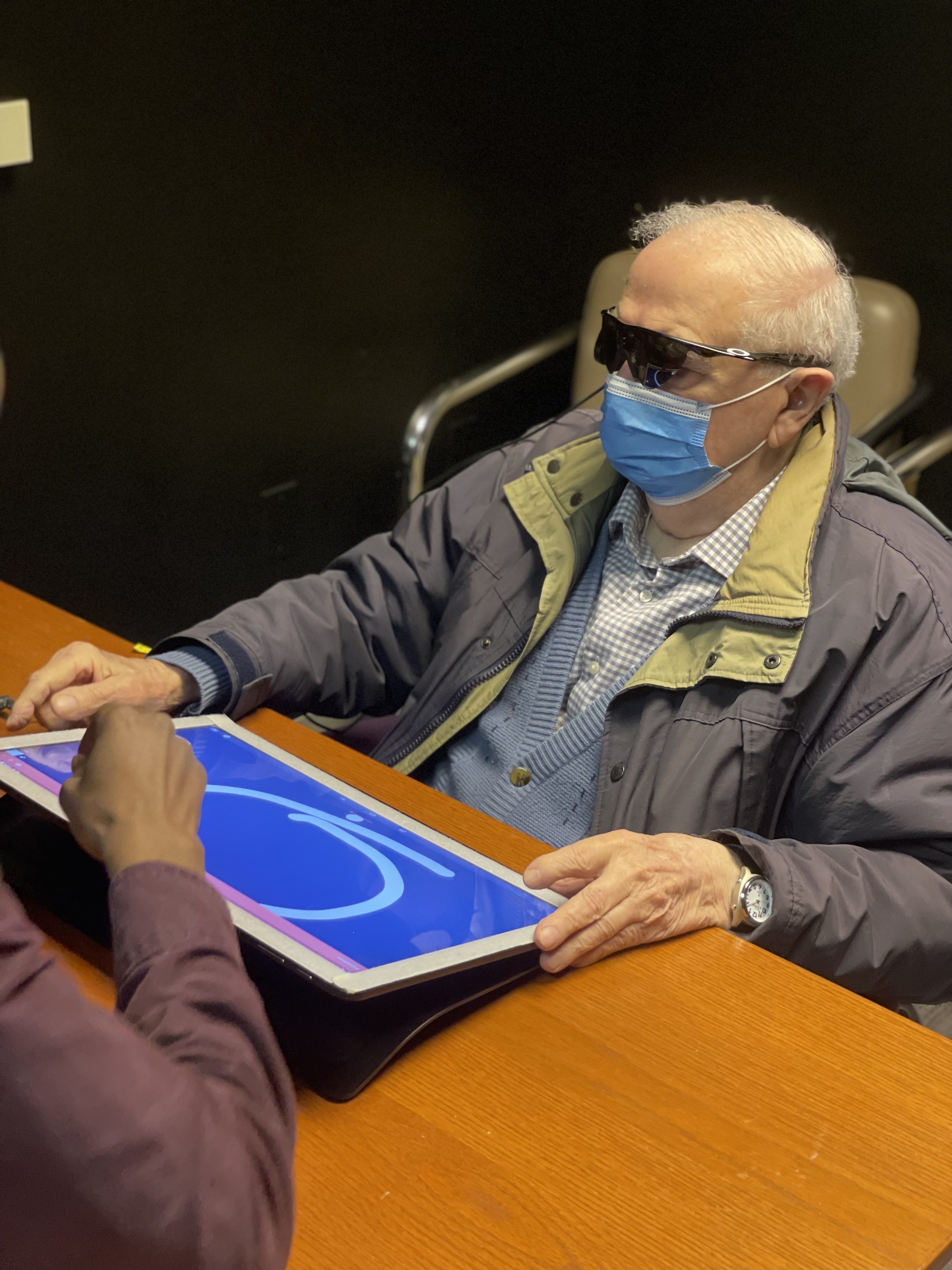 An Argus II user wearing the system’s camera glasses and a face mask sits at a table with a touchscreen showing a traced curved line; researcher Byron Johnson sits across the table.