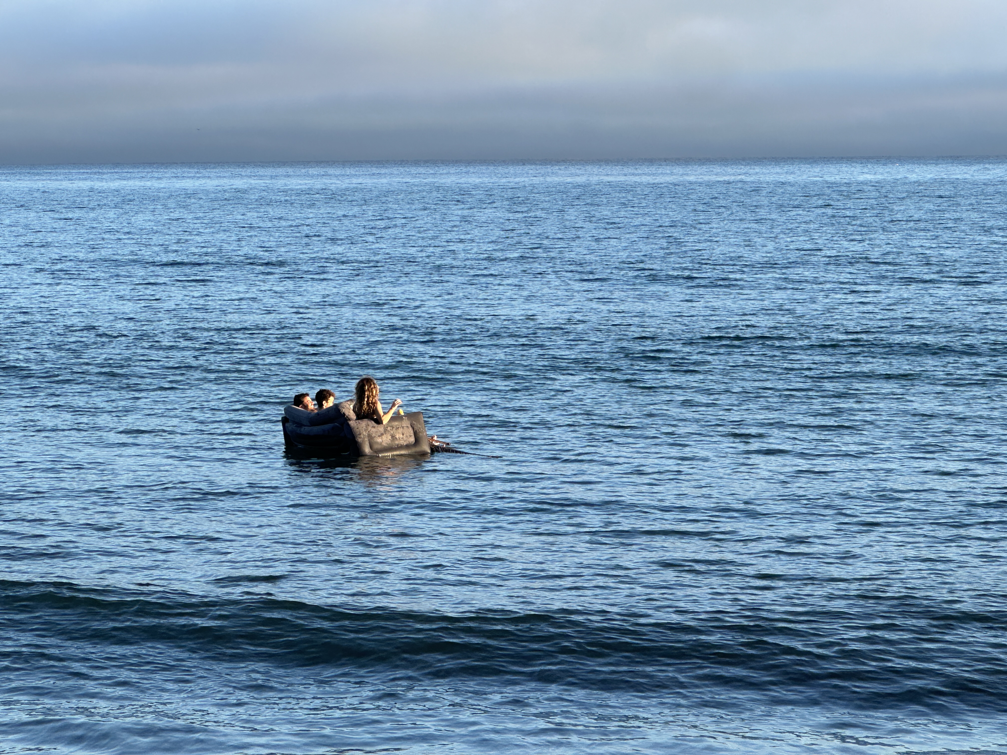 Three people float on a sofa in calm ocean water under an overcast evening sky.
