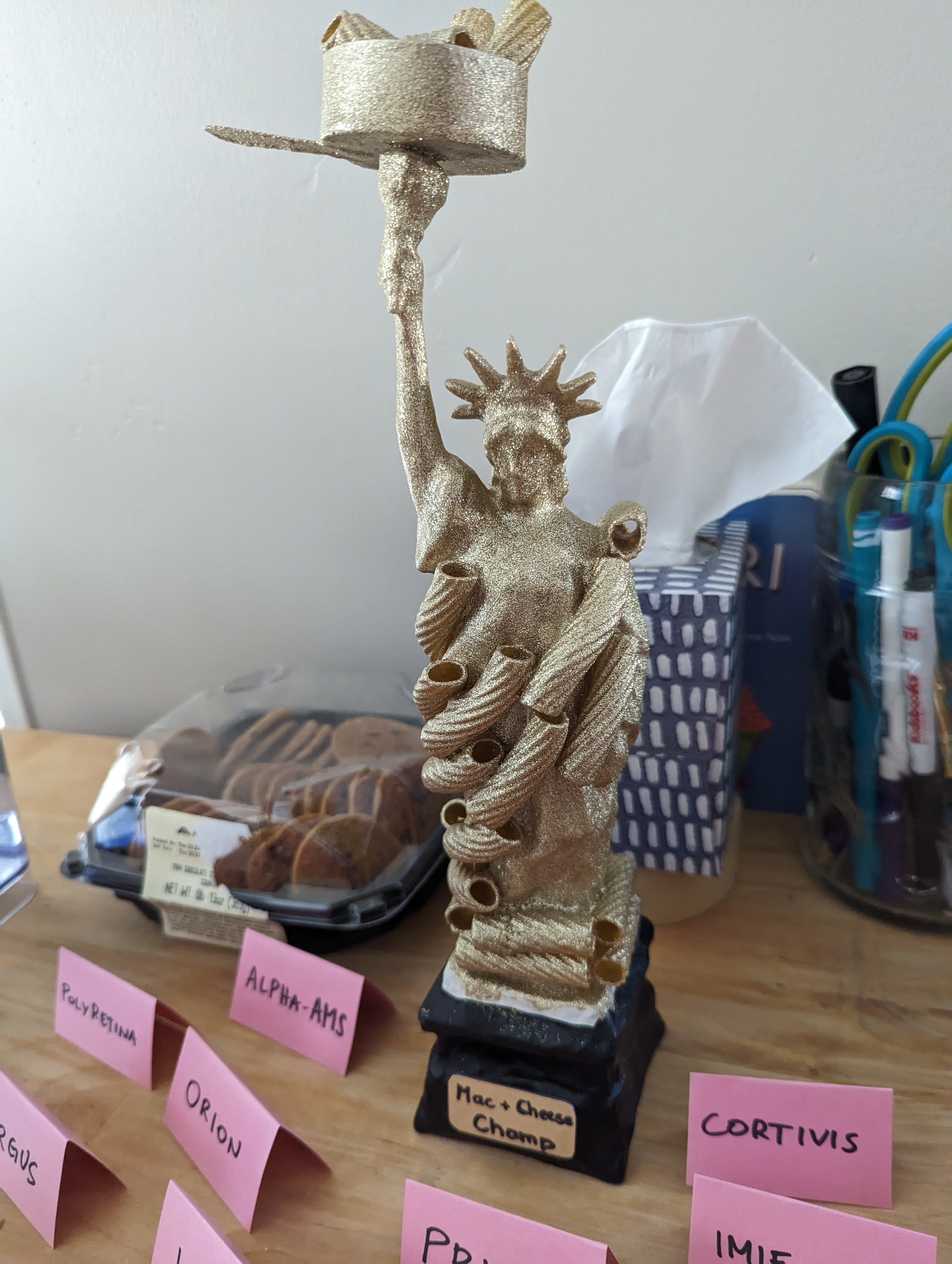 Gold-glitter Statue of Liberty figurine covered in macaroni, holding up a saucepan instead of a torch, on a base labeled “Mac + Cheese Champ.” Tent cards around it standing for the anonymous competition entries are named after visual prostheses: PolyRetina, Alpha-AMS, Orion, Argus, and Cortivis.