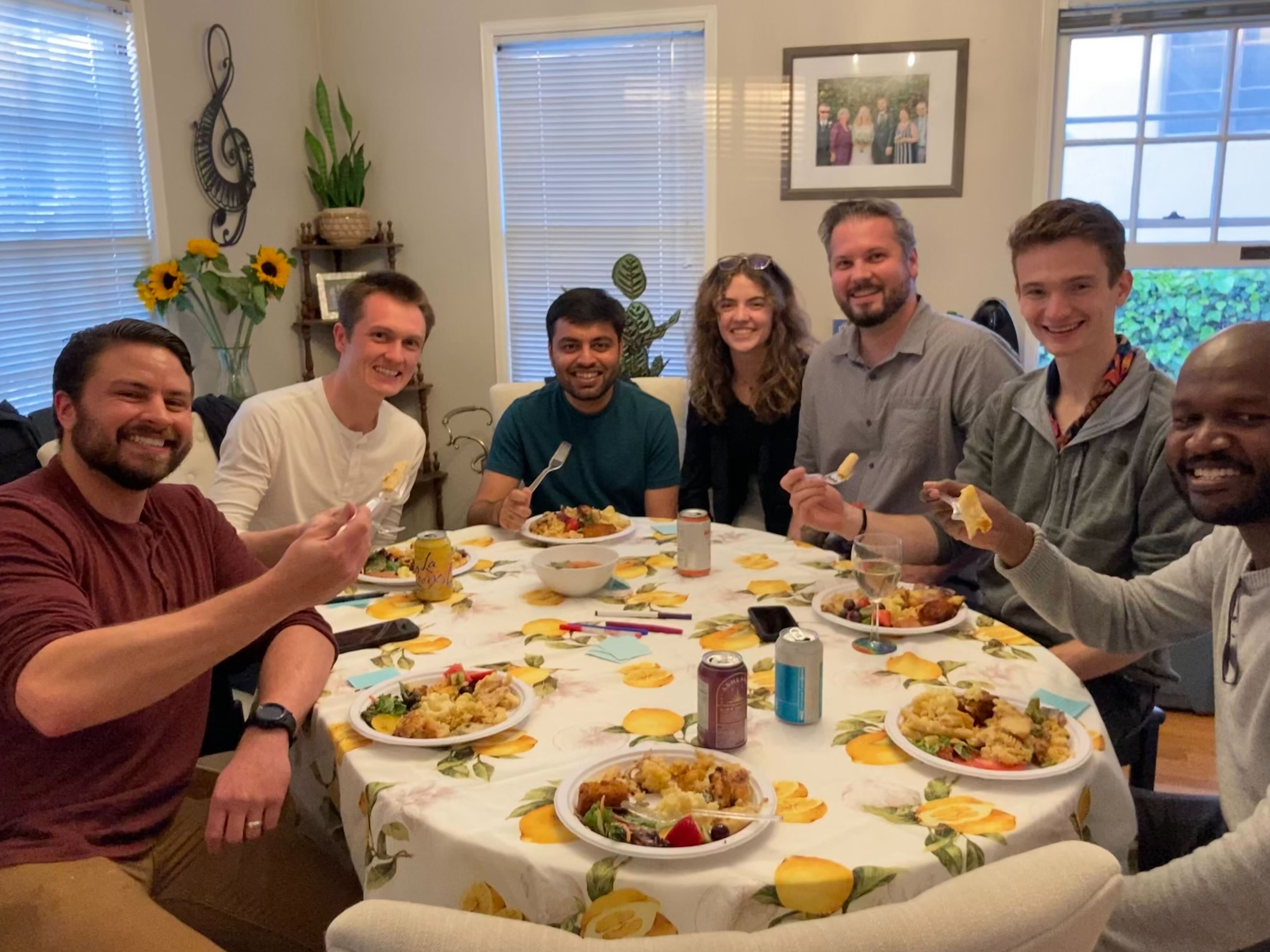Seven lab members smile around a round dining table with a lemon-print tablecloth and plates of mac and cheese, several raising forks (ca. 2022). From left to right: Justin Kasowski, Galen Pogoncheff, Apurv Varshney, Lily Turkstra, Prof. Beyeler, Jacob Granley, and Byron Johnson.