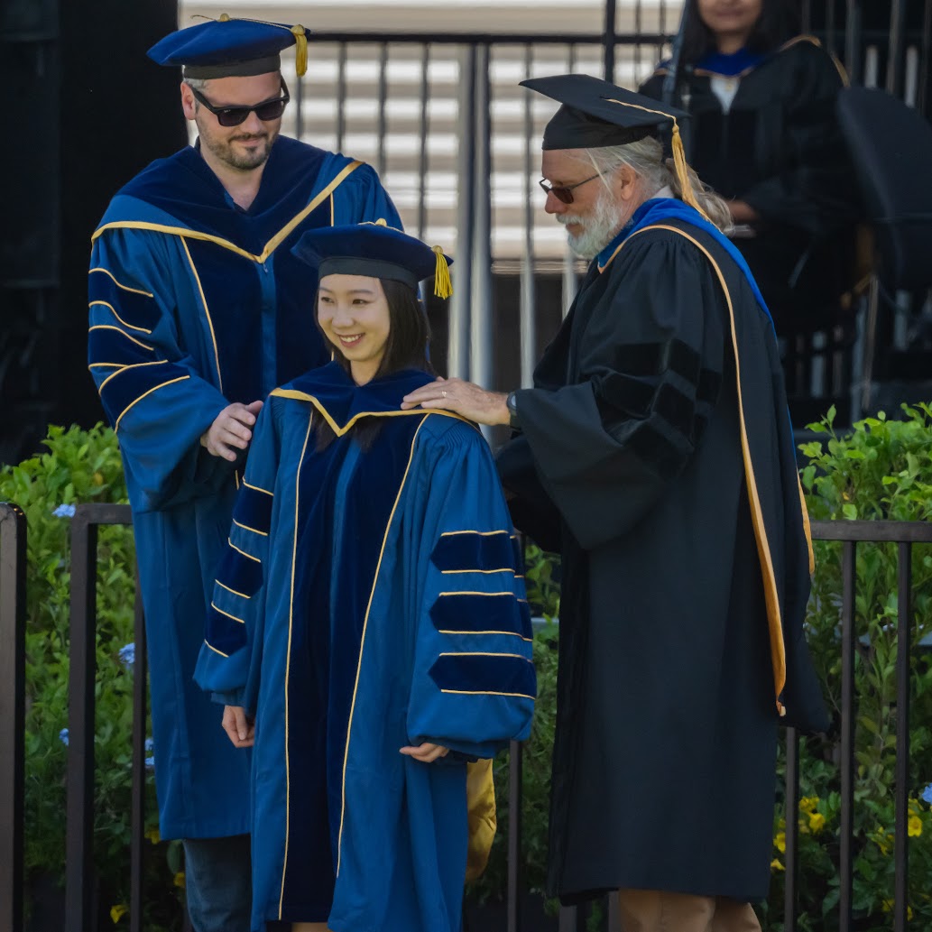 Aiwen Xu, in blue doctoral robes, smiles as two faculty members place the doctoral hood.